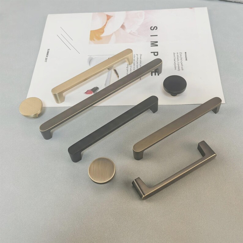May include: A selection of cabinet handles and knobs in gold, bronze, and black finishes. The image shows a variety of bar pulls and round knobs. The word "SIMPLE" is visible on a magazine page. The hardware is displayed on a neutral background.