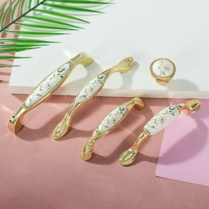 Pu&ograve; includere: Un set di ferramenta dorata per armadi, tra cui maniglie e un pomello. Ogni pezzo presenta un centro in ceramica bianca con un delicato motivo floreale in blu, rosa e verde. La ferramenta &egrave; esposta su una superficie rosa e bianca.