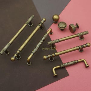 Può includere: Una collezione di ferramenta per mobili in ottone antico, tra cui vari stili di maniglie e pomelli. Le maniglie presentano design a coste, strutturati e a motivi. L'hardware è esposto su uno sfondo marrone e rosa.