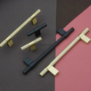 May include: An assortment of cabinet pulls in gold and black. The gold pulls have a textured, diamond-patterned handle and a rectangular base. The black pulls have a similar diamond pattern. The pulls are arranged on a brown and pink background.