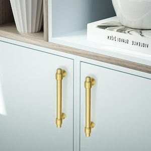 Peut inclure: Gros plan sur deux portes d'armoires bleu clair avec des poign&eacute;es verticales dor&eacute;es. Les poign&eacute;es sont cylindriques avec des d&eacute;tails d&eacute;coratifs. Les portes de l'armoire font partie d'un meuble plus grand, avec une &eacute;tag&egrave;re blanche et un vase d&eacute;coratif visible.