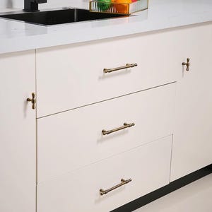 May include: Cream kitchen cabinets with antique brass handles. The drawers feature horizontal bar pulls, and the cabinet door has a T-shaped knob. The hardware provides a vintage aesthetic to the modern kitchen design.