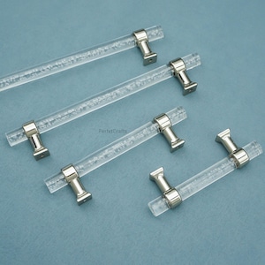 Peut inclure: Poign&eacute;es de meubles en acrylique transparent avec des fixations en m&eacute;tal argent&eacute;. Les poign&eacute;es sont dispos&eacute;es sur une surface bleu clair. Les tiges en acrylique ont un aspect textur&eacute;, semblable &agrave; des bulles. Les fixations sont rectangulaires avec un dessus arrondi.