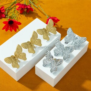 May include: Butterfly-shaped cabinet knobs in gold and silver. The knobs are displayed on white platforms against a warm orange background, with red flowers in the top left corner. The butterfly knobs are a decorative hardware item.