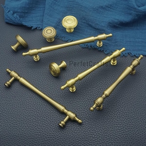 Puede incluir: Un surtido de herrajes de gabinete de latón antiguo, que incluyen perillas y tiradores. El hardware presenta un diseño clásico con detalles. Las piezas están dispuestas sobre una superficie azul oscuro, con un paño azul en el fondo. El texto "PerfecCrafts" es visible.