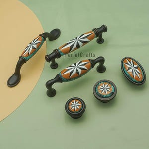 Może przedstawiać: Zestaw dekoracyjnych okuć do szafek, w tym uchwytów i gałek. Okucia mają wzór rozbłysku gwiazdy w kolorze białym, pomarańczowym i turkusowym, na tle ciemnego brązu. Widoczny jest tekst "PerfetcCrafts".