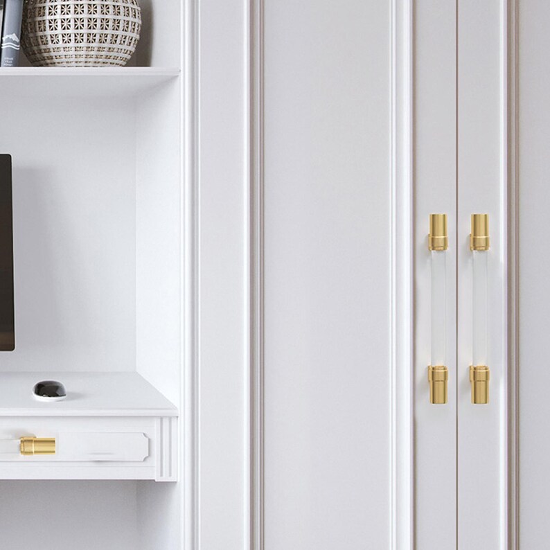 May include: White cabinet doors with clear acrylic and gold-toned hardware. The cabinet doors have a vertical panel design. The hardware is rectangular and mounted vertically. The cabinet is part of a white built-in unit.