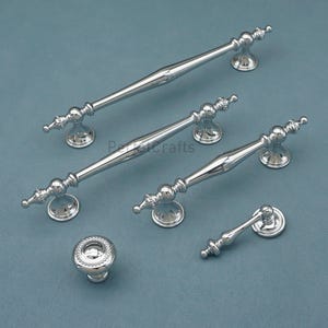 Może przedstawiać: Kolekcja polerowanych srebrnych okuć do szafek, w tym uchwyt&oacute;w i gałki. Uchwyty r&oacute;żnią się rozmiarem i wzornictwem, z dekoracyjnymi detalami. Okucia są prezentowane na niebiesko-szarej powierzchni.