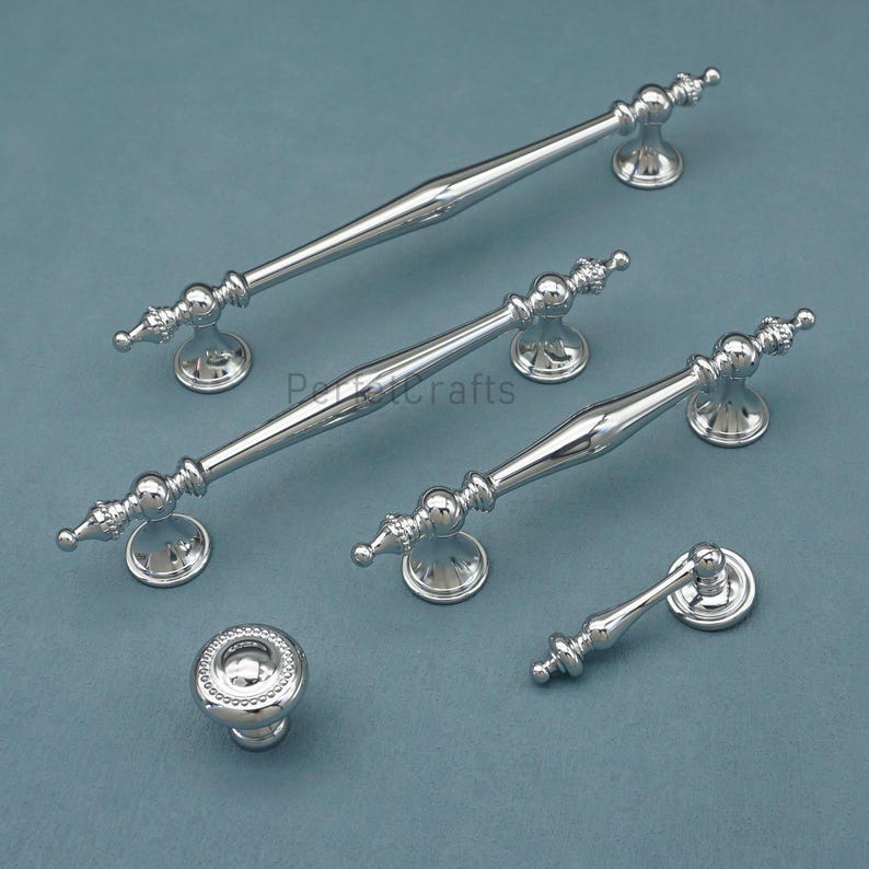 Może przedstawiać: Kolekcja polerowanych srebrnych okuć do szafek, w tym uchwyt&oacute;w i gałki. Uchwyty r&oacute;żnią się rozmiarem i wzornictwem, z dekoracyjnymi detalami. Okucia są prezentowane na niebiesko-szarej powierzchni.