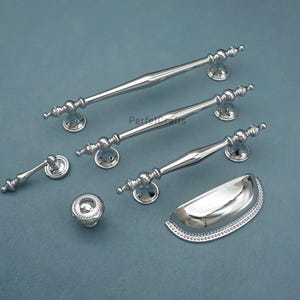 Może przedstawiać: Kolekcja polerowanych srebrnych okuć do szafek, w tym długie uchwyty prętowe, uchwyt miseczkowy i okrągła gałka. Okucia mają dekoracyjne detale i są prezentowane na niebiesko-szarej powierzchni.