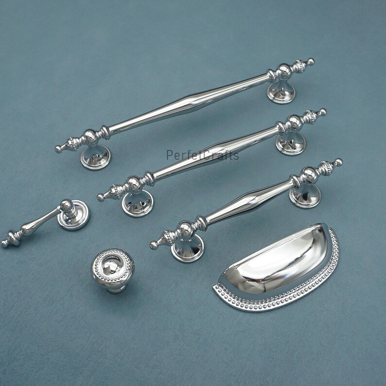 Może przedstawiać: Kolekcja polerowanych srebrnych okuć do szafek, w tym długie uchwyty prętowe, uchwyt miseczkowy i okrągła gałka. Okucia mają dekoracyjne detale i są prezentowane na niebiesko-szarej powierzchni.