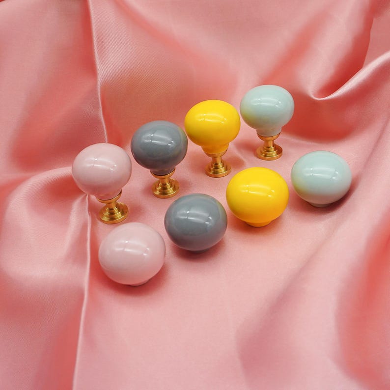 May include: Eight decorative cabinet knobs in a range of colours: pink, grey, yellow, and light blue. Each knob has a round, glossy ceramic top and a gold-coloured metal base. The knobs are displayed on a pink satin fabric.