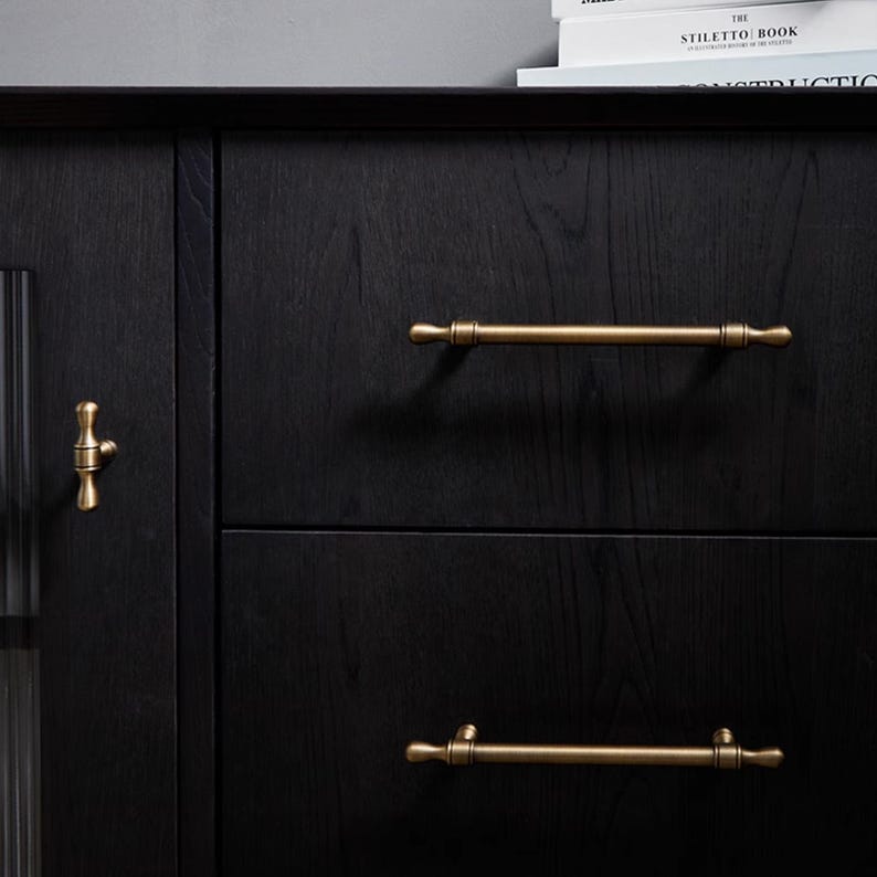 May include: A black wooden cabinet with brass handles and a door knob. The cabinet has two drawers with long bar handles and a door with a small knob. The wood grain is visible. The cabinet is likely for storage.