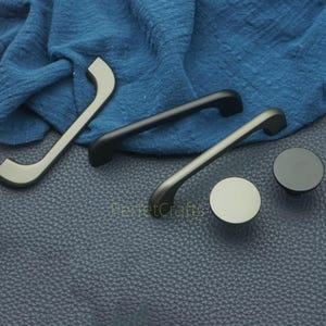 Op de afbeelding: Een selectie van kastgrepen en knoppen in verschillende metalen afwerkingen. De afbeelding toont zilveren, zwarte en grijze hardware. De handgrepen zijn gebogen of staafvormig en de knoppen zijn rond. De hardware wordt gepresenteerd op een getextureerd oppervlak.