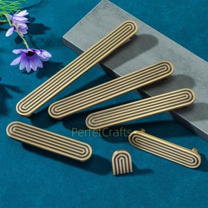 May include: A collection of brass-toned cabinet pulls and knobs with a grooved design. The hardware is arranged on a teal surface with a gray block and purple flowers. The pulls vary in size, with the longest being approximately 8 inches.