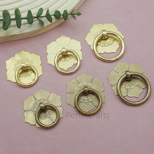 May include: Six brass flower-shaped cabinet pulls with ring handles. The pulls are in various shades of gold and bronze, with a floral design. The pulls are arranged on a pink surface.