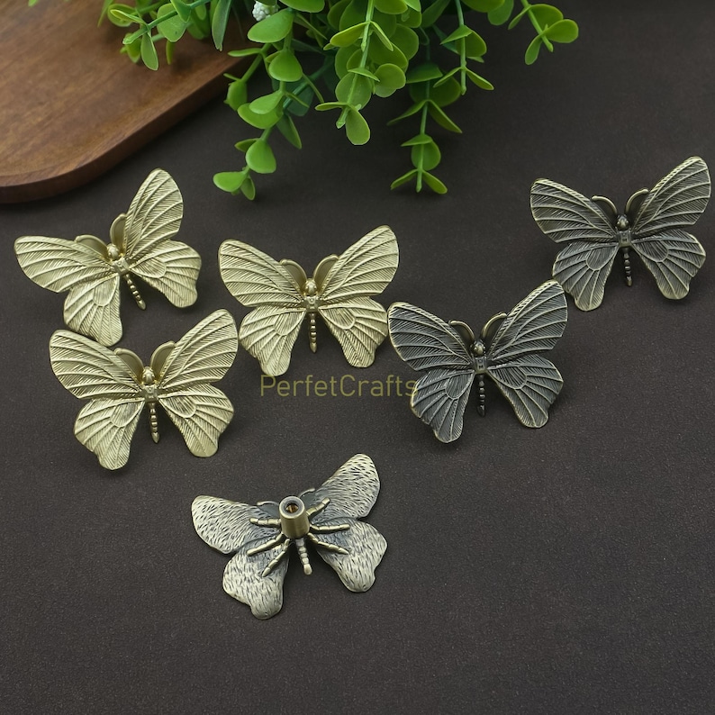 May include: A collection of decorative butterfly-shaped hardware in various metallic finishes. The butterflies are detailed with textured wings and are likely used as drawer pulls or cabinet knobs. The image includes the text "PerfetcCrafts".