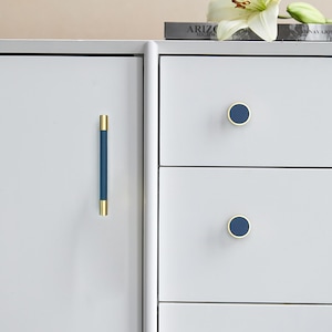 Puede incluir: Primer plano de un armario blanco con tiradores y pomos azules y dorados. El tirador es una barra vertical con centro azul y extremos dorados. Los pomos son redondos con centro azul y borde dorado.