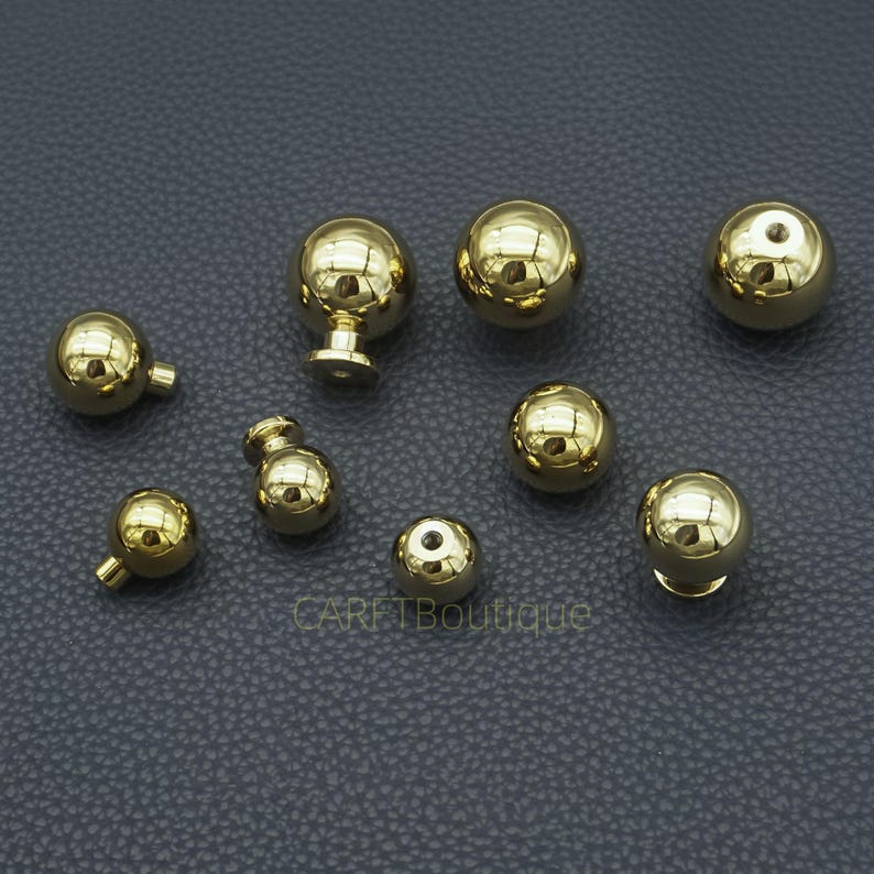 Puede incluir: Una colecci&oacute;n de tiradores y pomos dorados y brillantes de varios tama&ntilde;os. Los herrajes tienen un acabado pulido y reflectante, algunos con forma de bola y otros con un v&aacute;stago para la fijaci&oacute;n. Los art&iacute;culos est&aacute;n dispuestos sobre una superficie gris oscuro.