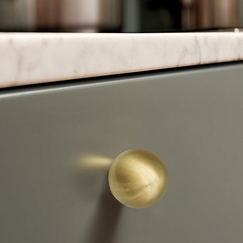 Op de afbeelding: Een close-up van een goudkleurige, bolvormige kastknop. De knop is bevestigd aan een grijze kastdeur. Een marmeren aanrechtblad is zichtbaar op de achtergrond.
