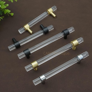 May include: Clear acrylic cabinet handles with gold, black, and silver accents. The handles are cylindrical with a T-bar design. Several handles are arranged on a brown surface. The text "CAPEtique" is visible on the surface.