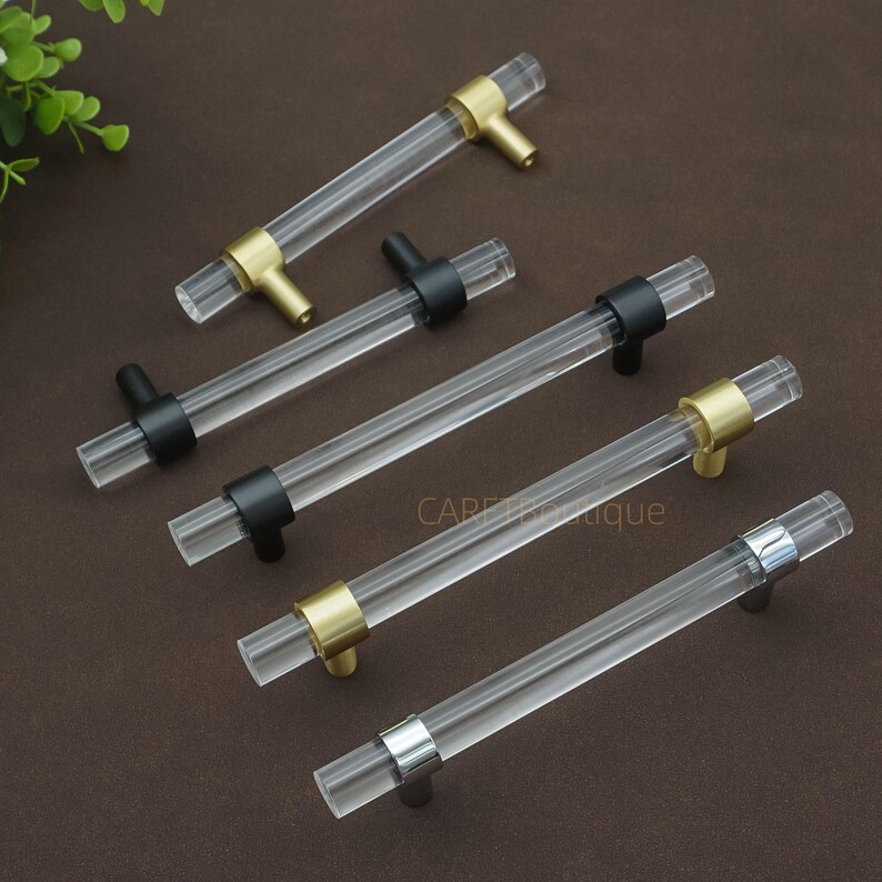 May include: Clear acrylic cabinet handles with gold, black, and silver accents. The handles are cylindrical with a T-bar design. Several handles are arranged on a brown surface. The text "CAPEtique" is visible on the surface.