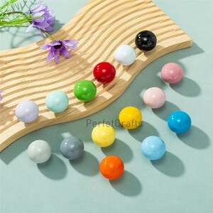 May include: A selection of multi-coloured, spherical beads, including red, green, yellow, and blue, displayed on a wooden, wave-shaped tray. The beads are scattered on a light blue surface, with some purple flowers in the top left corner.