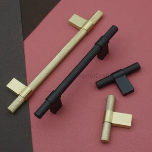 K&ouml;nnte beinhalten: Eine Auswahl an Schrankgriffen, darunter goldene und schwarze strukturierte Stangengriffe und ein T-f&ouml;rmiger Griff. Die goldenen Griffe haben eine quadratische Basis, w&auml;hrend die schwarzen Griffe eine rechteckige Basis haben. Pr&auml;sentiert auf einer rosa und braunen Oberfl&auml;che.