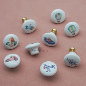 以下が含まれることがあります： 金色のベースが付いた白いセラミック製のキャビネットノブ。各ノブには、車、飛行機、ボート、ヘリコプター、熱気球など、さまざまな輸送手段の水彩イラストが描かれています。ノブは家具用に設計されています。