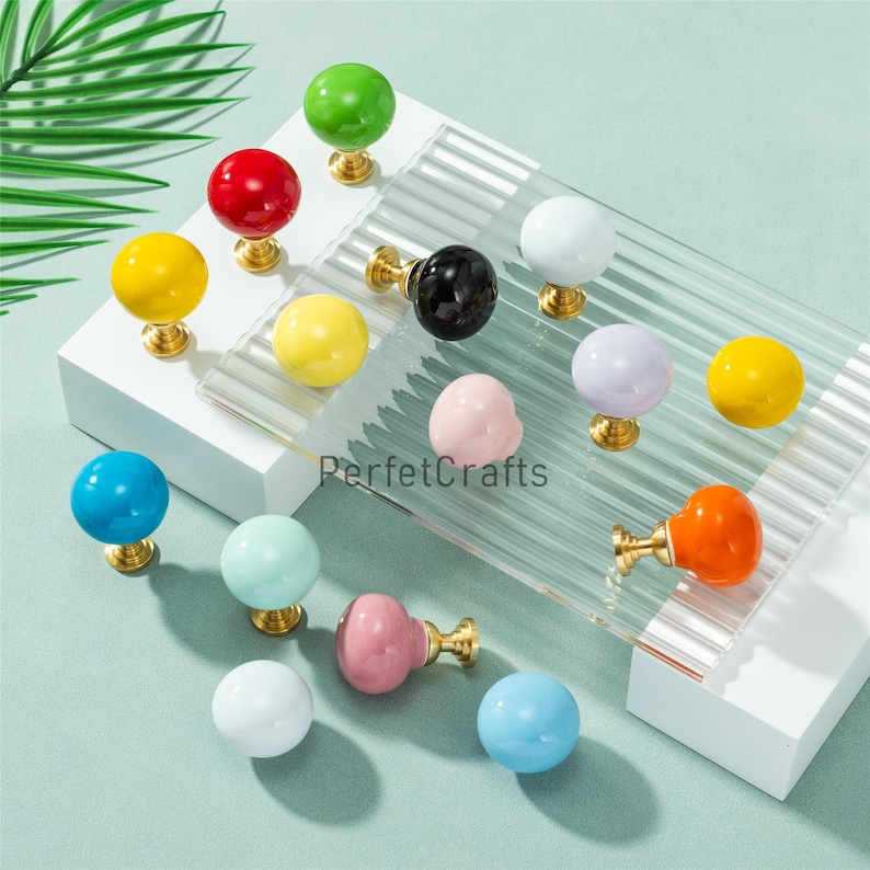 May include: A selection of colourful cabinet knobs in shades like red, yellow, green, blue, and pink. Each knob has a spherical form and a gold-coloured base. The knobs are displayed on a clear, textured surface.