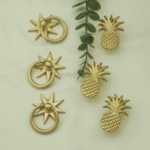 May include: Gold-coloured cabinet hardware featuring starburst ring pulls and pineapple-shaped knobs. The hardware is arranged on a light background with a sprig of greenery.