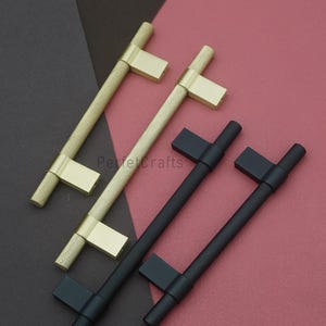 K&ouml;nnte beinhalten: Schrankgriffe in Gold und Schwarz. Die goldenen Griffe haben einen strukturierten, rautenf&ouml;rmigen Griff und eine rechteckige Basis. Die schwarzen Griffe haben einen &auml;hnlichen strukturierten Griff und eine Basis. Auf einer rotbraunen Oberfl&auml;che pr&auml;sentiert.