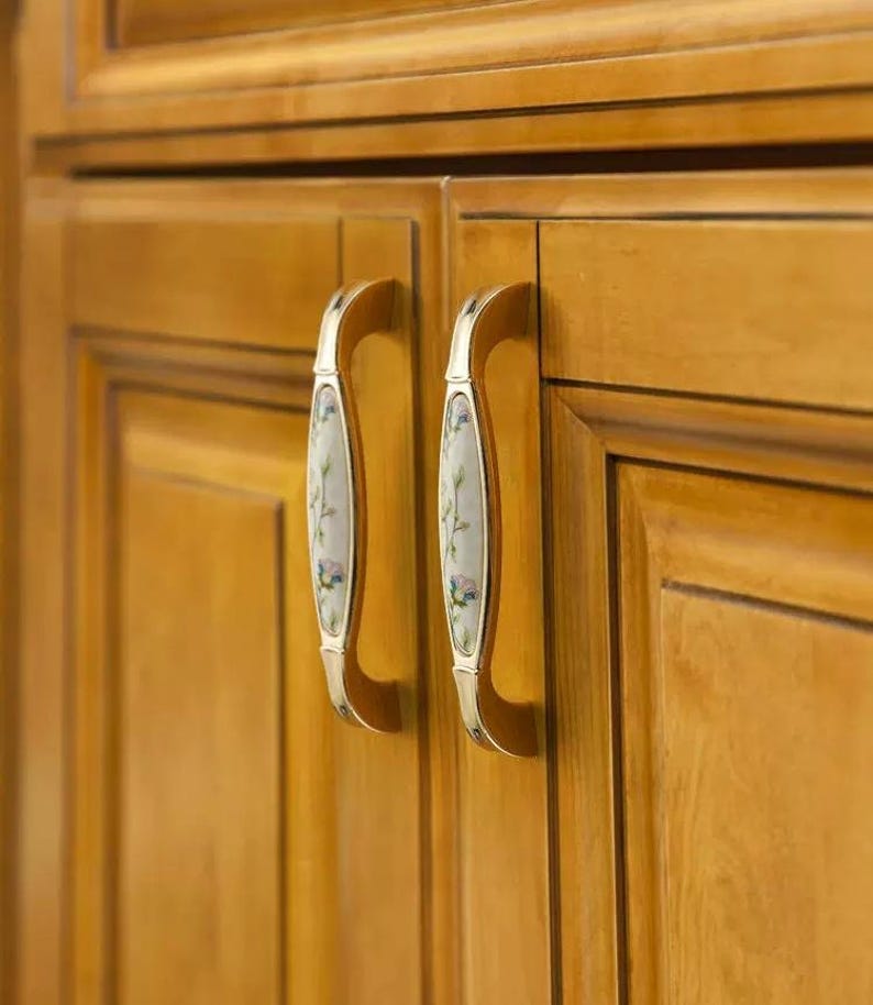 Pu&ograve; includere: Primo piano di due ante di armadi con ferramenta decorativa. Le ante dell'armadio sono in legno marrone chiaro. Ogni anta ha una maniglia curva con un motivo floreale su sfondo bianco e dettagli metallici color oro.