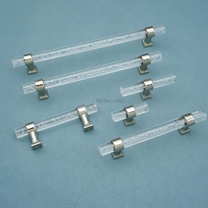 Peut inclure: Poignées de meubles en acrylique transparent avec bases en métal argenté. Les poignées présentent une texture de type bulle à l'intérieur de l'acrylique transparent. Plusieurs poignées de différentes tailles sont présentées sur fond bleu.