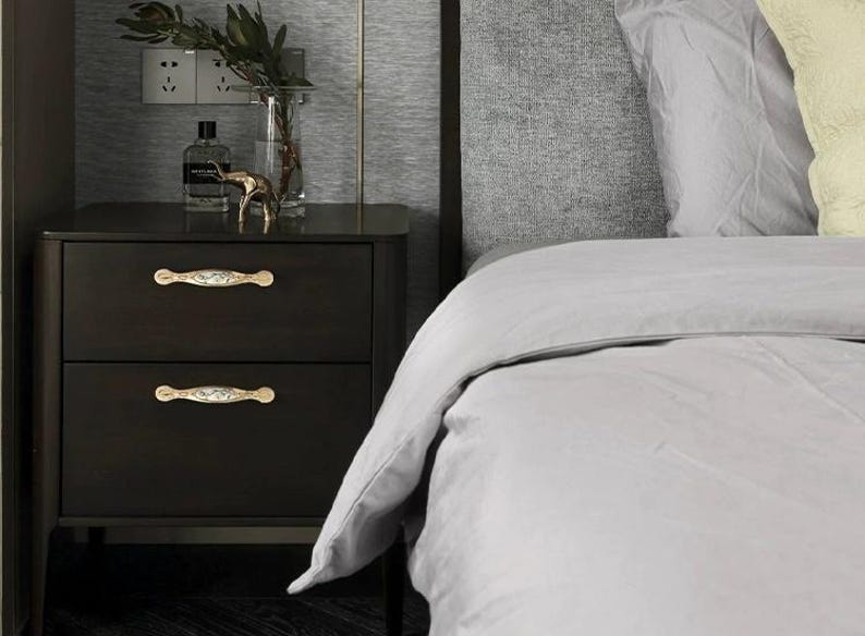 Pu&ograve; includere: Comodino marrone scuro con due cassetti, con maniglie decorative color oro. Un vaso di vetro con verde, una bottiglia di profumo e una piccola statuina d'oro sono in cima. Il comodino &egrave; accanto a un letto con biancheria da letto grigia.