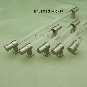 May include: Brushed nickel cabinet pulls with clear acrylic rods. The hardware has a modern design with a cylindrical shape and square base. The text "Brushed Nickel" and "PerfecCrafts" are visible.