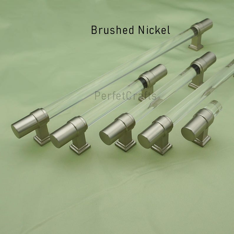 May include: Brushed nickel cabinet pulls with clear acrylic rods. The hardware has a modern design with a cylindrical shape and square base. The text "Brushed Nickel" and "PerfecCrafts" are visible.