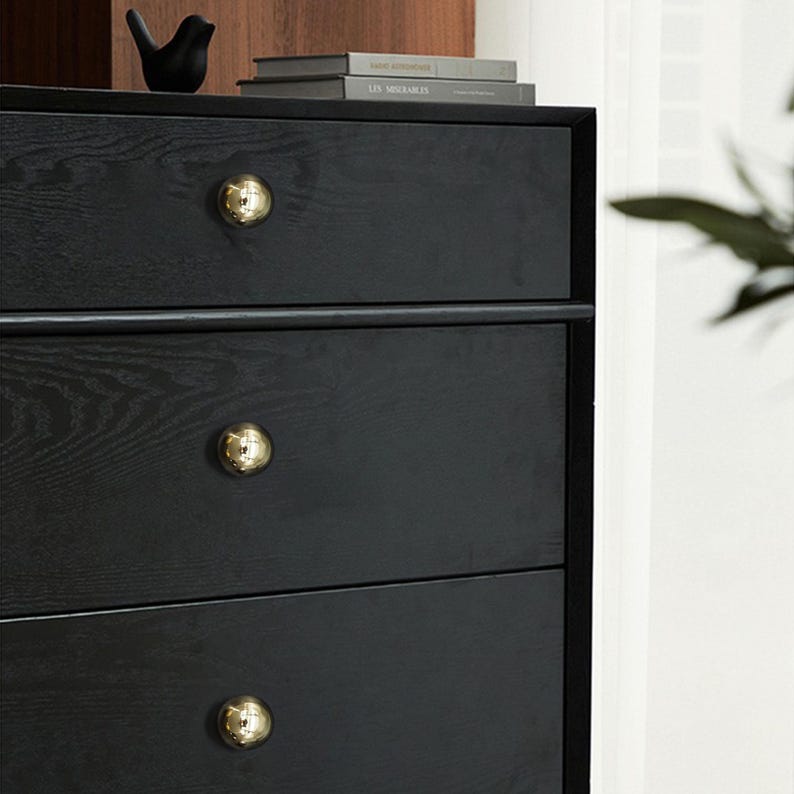 Op de afbeelding: Close-up van een zwarte ladekast met drie lades, elk versierd met een gouden bolvormige knop. De houtnerf is zichtbaar. Een klein zwart vogelbeeldje en boeken staan bovenop de kast.