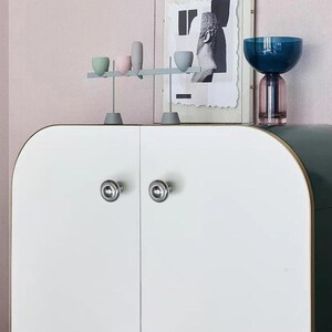 Może przedstawiać: Biała szafka z dwoma srebrnymi gałkami, ozdobiona elementami dekoracyjnymi. Niebieski i r&oacute;żowy szklany wazon znajduje się po prawej stronie, a po lewej stronie znajduje się ekspozycja małych, kolorowych ceramicznych element&oacute;w. W tle oprawiona czarno-biała grafika.