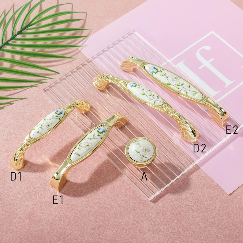 Pu&ograve; includere: Una collezione di ferramenta per mobili color oro, tra cui maniglie e un pomello. La ferramenta presenta un intarsio in ceramica bianca con un design floreale nei toni del blu, rosa e verde. Le maniglie variano per dimensioni e forma, con dettagli ornati.