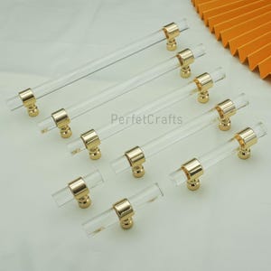 May include: Clear acrylic cabinet pulls with gold-toned hardware. The pulls are cylindrical and have a modern design. The hardware is a shiny gold color. The pulls are arranged diagonally on a white surface.