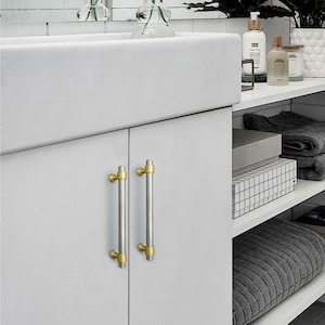 Peut inclure: Deux poign&eacute;es de meuble avec une finition argent bross&eacute; et des accents dor&eacute;s. Les poign&eacute;es cylindriques ont des embouts dor&eacute;s &agrave; chaque extr&eacute;mit&eacute; et sont mont&eacute;es sur une porte d'armoire blanche. La quincaillerie ajoute une touche d'&eacute;l&eacute;gance &agrave; la salle de bain.