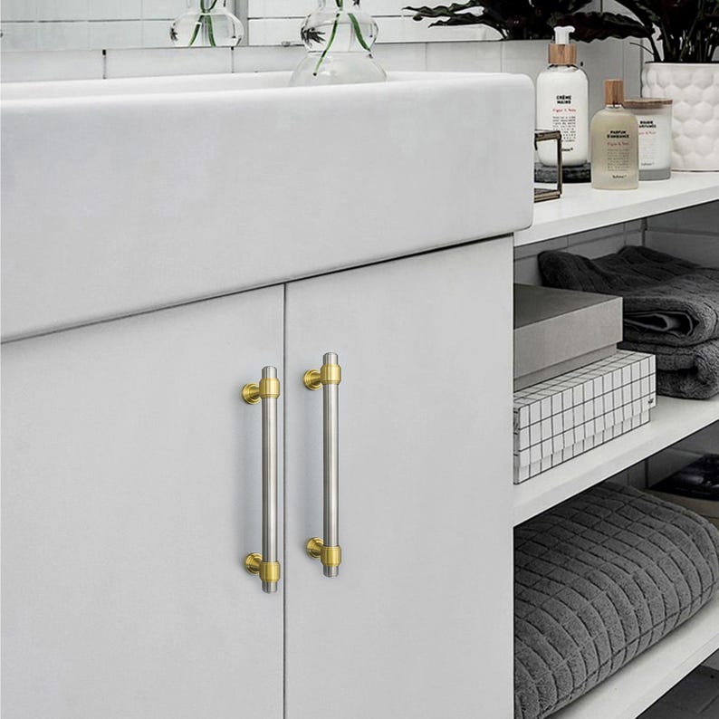 Peut inclure: Deux poign&eacute;es de meuble avec une finition argent bross&eacute; et des accents dor&eacute;s. Les poign&eacute;es cylindriques ont des embouts dor&eacute;s &agrave; chaque extr&eacute;mit&eacute; et sont mont&eacute;es sur une porte d'armoire blanche. La quincaillerie ajoute une touche d'&eacute;l&eacute;gance &agrave; la salle de bain.
