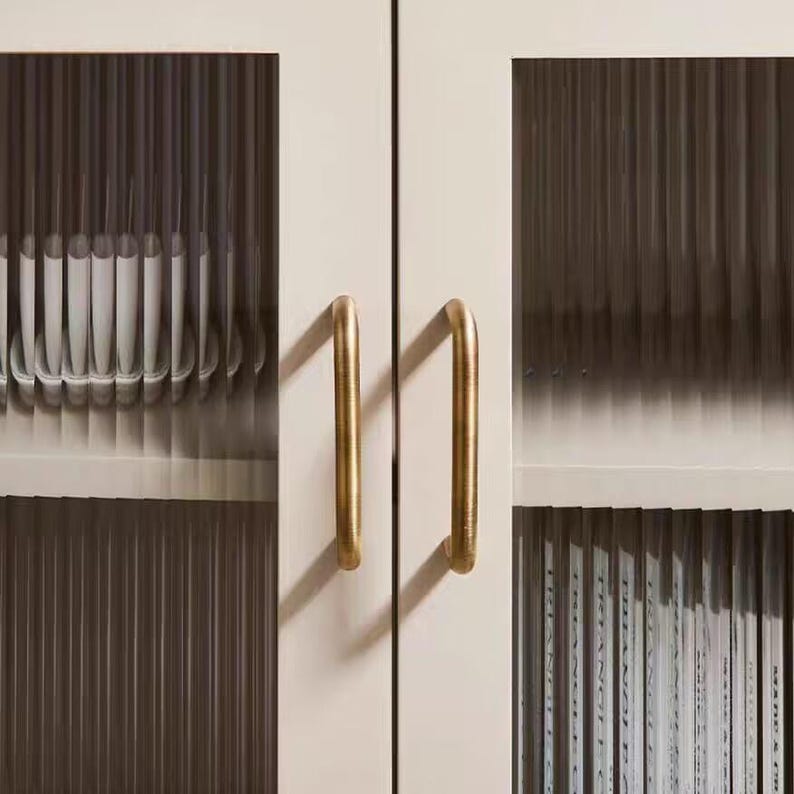 K&ouml;nnte beinhalten: Nahaufnahme eines cremefarbenen Schranks mit zwei T&uuml;ren und goldfarbenen Griffen. Die Schrankt&uuml;ren haben Glaspaneele mit vertikalen Rippendetails. Der Schrank ist mit wei&szlig;en Objekten und Zeitschriften gef&uuml;llt.