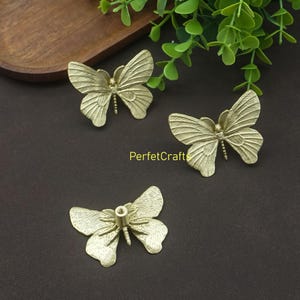 May include: Three brass butterfly-shaped cabinet knobs. The knobs have detailed wing patterns and a central screw post. The knobs are arranged on a dark brown surface with green foliage in the background. The text "PerfetcCrafts" is visible.