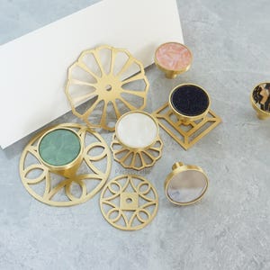 Puede incluir: Una colección de pomos decorativos para armarios en varios diseños y colores. Los pomos presentan bases de metal dorado con tapas en rosa, negro, blanco y verde. Algunos tienen patrones geométricos, otros diseños circulares o florales.