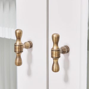 May include: Close-up of two antique brass cabinet pulls mounted on a white cabinet door. The pulls have a decorative, ornate design with a vintage aesthetic. The hardware is a warm, metallic colour.