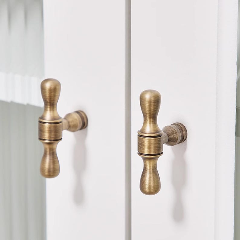 May include: Close-up of two antique brass cabinet pulls mounted on a white cabinet door. The pulls have a decorative, ornate design with a vintage aesthetic. The hardware is a warm, metallic colour.
