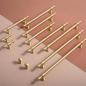 May include: A selection of gold-toned cabinet hardware, comprising long bar pulls and smaller knobs. The pulls feature a cylindrical bar with decorative end pieces. The knobs are rounded with a matching gold finish. The hardware is displayed on a two-tone pink and brown surface.