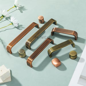 Może przedstawiać: Asortyment mosiężnych i brązowych uchwytów do szafek, w tym uchwyty i gałki. Uchwyty mają zakrzywiony kształt z wieloma równoległymi liniami. Gałki są cylindryczne i zaokrąglone. Okucia są ułożone na jasnoniebieskiej powierzchni.